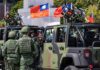 Soldiers in camouflage uniforms standing near military vehicles with flags in the background