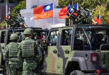 Soldiers in camouflage uniforms standing near military vehicles with flags in the background