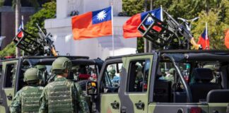 Soldiers in camouflage uniforms standing near military vehicles with flags in the background
