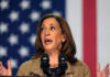 A woman speaking passionately at a podium with microphones against an American flag backdrop