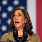 A woman speaking passionately at a podium with microphones against an American flag backdrop