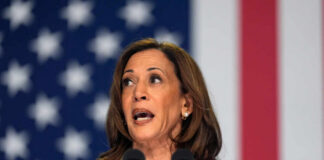 A woman speaking passionately at a podium with microphones against an American flag backdrop