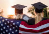 Two graduates wearing caps holding an American flag during sunset