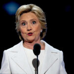 Legacy Bombshell: Clinton Turns On Biden A woman in a white suit speaking at a podium during a conference