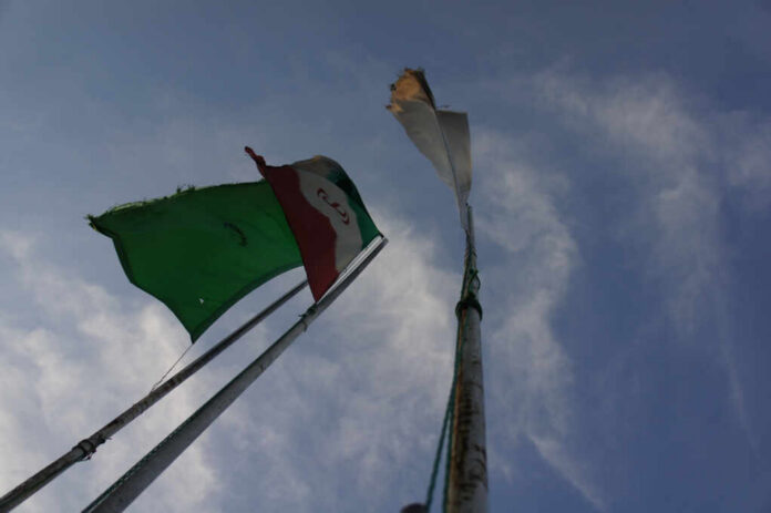 Iran's,Flag,With,The,Background,Of,Sky,And,A,Green Two flags on a flagpole against a blue sky with clouds
