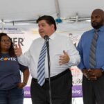 Trump Dares Pritzker: Call For Help man speaking at a podium with three people standing beside him