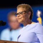 Bass Family Pulled Into Inferno Fight A woman in a light blue dress speaking at a podium with a microphone