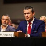 A man in a blue suit and red tie sitting at a hearing table with a nameplate