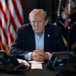 Capitol Meltdown Over Trump’s Hardball Linkage A serious-looking man seated at a table with phones and an American flag in the background