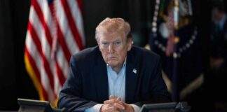 Capitol Meltdown Over Trump’s Hardball Linkage A serious-looking man seated at a table with phones and an American flag in the background