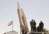Hezbollah Cheers ‘Victory’—What Did Washington Trade? Military personnel standing near missile launchers with an Iranian flag in the background