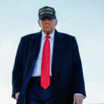 A political figure wearing a black coat and red tie, with a Make America Great Again hat, walking outdoors