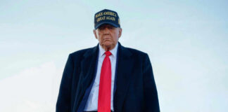 A political figure wearing a black coat and red tie, with a Make America Great Again hat, walking outdoors