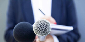 A person holding two microphones, one black and one silver, with a notebook in the background