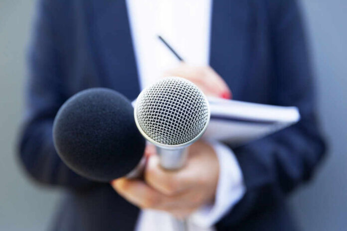 shutterstock_1480323476 A person holding two microphones, one black and one silver, with a notebook in the background
