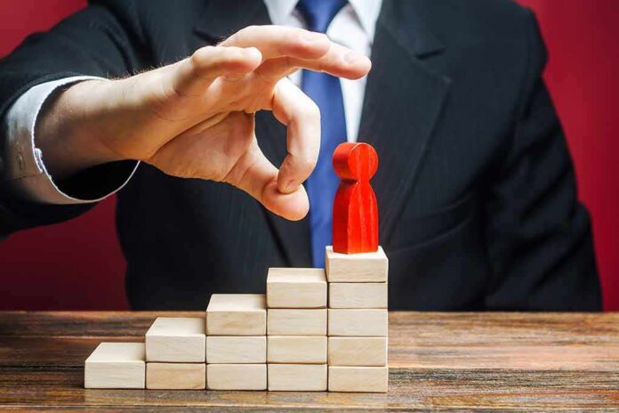shutterstock_1613353153 Businessperson hand above red figure on wooden block steps