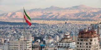Obliterate Iran? Carolina Firebrands Diverge Iranian flag over a dense cityscape with mountains in the background