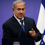man in suit speaking at podium with Israeli flag