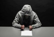 Person in gray hoodie writing while handcuffed at a table