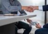 Two people shake hands over a desk while cash is exchanged underneath