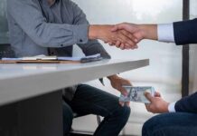 Two people shake hands over a desk while cash is exchanged underneath