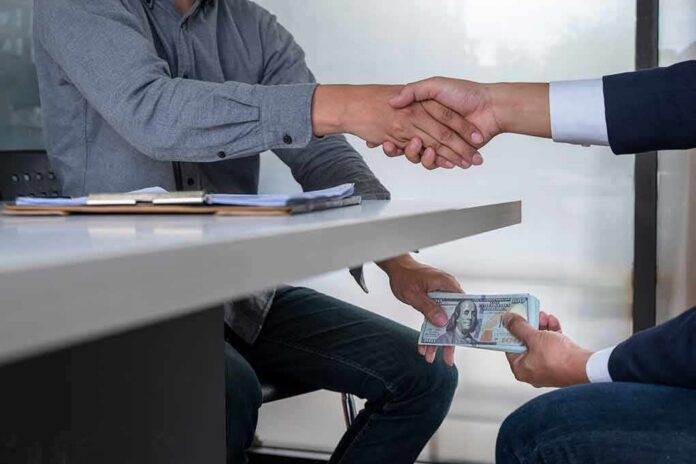 Two people shake hands over a desk while cash is exchanged underneath