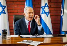 man in suit speaking at a desk with Israeli flags behind him
