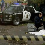 Mass Grave Found At ‘Rescue’ Crime scene with police car and two investigators by a covered body