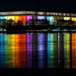 Court FREEZES Shutdown, Name Rollback Ordered Modern building lit in rainbow colors reflected on water at night
