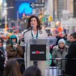 Kathy Hochul speaks at a clear podium in a city street; Josh Boyce stands among the crowd