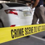 Yellow crime scene tape in front of a white car with investigators nearby