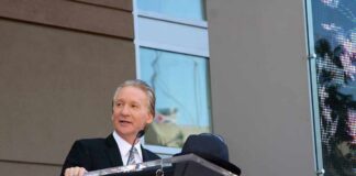 Bill Maher speaks at a podium during an outdoor event