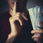 Person holding US dollar bills while making a shushing gesture