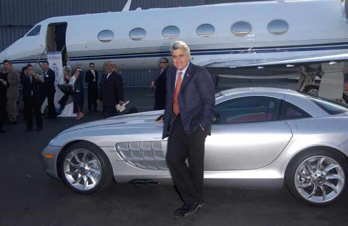 Jay Leno leaning on a silver sports car near a private jet