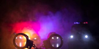 Police Punch Caught – Bodycam Gaps Fuel Big Questions Handcuffs on a surface with police cars and flashing lights in smoky darkness