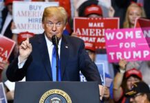 Donald Trump speaking at a rally podium with crowd holding signs