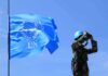 Winter Blackouts – War Crimes Alleged UN peacekeeper with blue helmet looks through binoculars beside UN flag