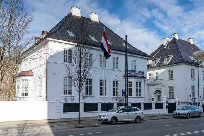 White embassy building with flag and parked cars on city street