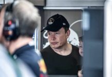 Elon Musk wearing a black cap in a crowded indoor setting
