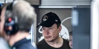 Elon Musk wearing a black cap in a crowded indoor setting