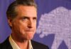 Newsom Picks Fight – Feds Won’t Blink Gavin Newsom at a press event with a purple backdrop