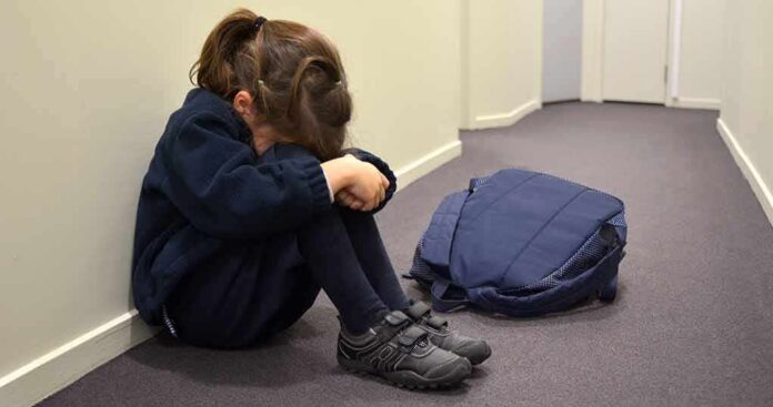 Child sitting on hallway floor with head down beside backpack